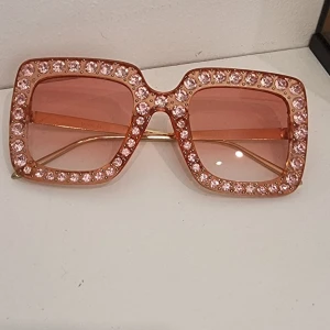 Rosa glittriga oversized solglasögon - Stora fyrkantiga solglasögon i rosa med glittriga stenar runt hela bågen. Snygg statement-accessoar med tonade rosa glas och tunna guldfärgade skalmar. Perfekt för dig som vill sticka ut och addera extra bling till din look.