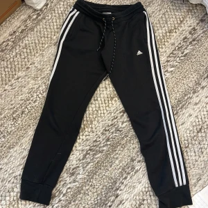 Svarta Adidas mjukisbyxor S - Svarta mjukisbyxor från Adidas med klassiska vita ränder längs sidorna och liten logga på låret. Byxorna har snörning i midjan och muddar vid bensluten. Storlek S men lite mindre i storlek. 