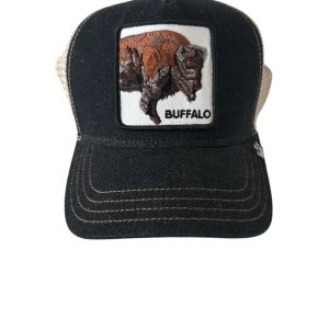 Svart och beige keps från The Farm - Cool keps med svart front och beige mesh baktill. Framsidan har en broderad patch med en buffel och texten 'BUFFALO'. Justerbar snapback och böjd skärm. Perfekt för dig som vill sticka ut med en unik accessoar.