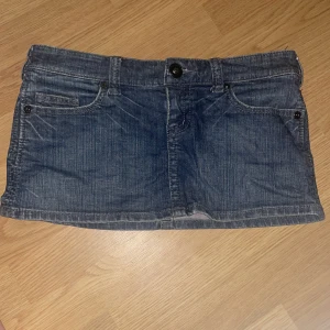 Jeanskjol från River Island med nitar - Snygg blå jeanskjol från River Island med klassisk femficksdesign och coola nitar på bakfickorna. Kort modell med knapp och dragkedja framtill. Storlek 10 typ S  priset kan diskuteras. Skriv om ni vill ha mer bilder 