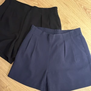 Svarta & marinblå shorts Gina Tricot - Två par stilrena shorts från Gina Tricot i storlek 36. Ena paret är svart och det andra är marinblått, båda med hög midja och diskreta veck framtill. Perfekta för en clean och enkel look. Materialet känns mjukt och har en lätt struktur.