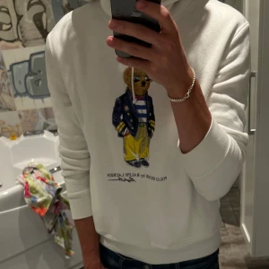 Vit hoodie Polo Bear Ralph Lauren - Vit hoodie från Ralph Lauren med ikoniska Polo Bear-trycket framtill. Tröjan har huva med snörning och ribbade muddar. Klassisk och clean look med en cool björn i preppy outfit på bröstet. Perfekt för dig som gillar streetstyle och märkeskläder.