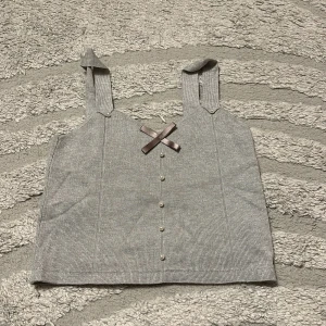 Grå ribbad linnetopp med rosett - Supersöt grå ribbad linnetopp med breda axelband och dekorativa pärlknappar framtill. Söt brun rosett framtill ger en extra detalj. Perfekt att matcha med jeans eller kjol för en chill look.