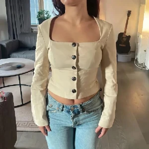 Beige croppad blus med knappar - Trendig beige blus med croppad passform och långa ärmar. Fyrkantig urringning och svarta knappar framtill ger en stilren look. Sitter figurnära och har en lätt strukturerad känsla i materialet. Perfekt till jeans för en snygg vardagsstil.