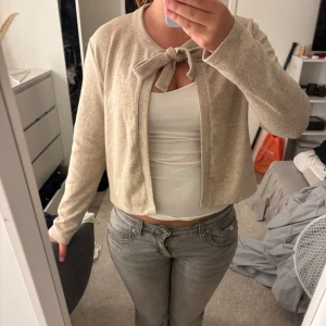 Beige kofta med knyt från 157 - Superfin beige kofta från 157 med knyt framtill vid halsen. Modellen är croppad och har långa ärmar. Mjuk och skön, perfekt för lager-på-lager. Passar dig som gillar en enkel men söt stil. Storlek S, pris kan diskuteras