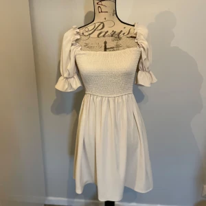 Ljus kortklänning med puffärmar - Supersöt ljus kortklänning med smockad överdel och puffiga kortärmar med volangkant. Klänningen har en fyrkantig ringning och ett flowy kjolparti som ger en romantisk vibe. Perfekt för varma dagar och somriga tillfällen.