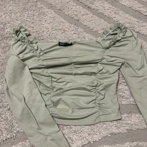Ljusgrön croppad topp från SHEIN - Trendig ljusgrön croppad topp från SHEIN med långa ärmar och snygga rynkade detaljer framtill och vid axlarna. Toppen har en offshoulder-design som ger en cool vibe och är perfekt att styla med höga jeans eller kjol.