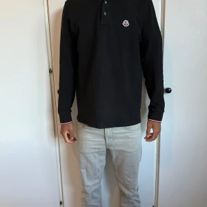 Svart pikétröja från Moncler - Svart långärmad pikétröja från Moncler med klassisk krage och knappar framtill. Tröjan har Moncler-logga på bröstet och snygga rödvita detaljer på krage och ärmslut. Tillverkad i bomull för en skön och stilren känsla.