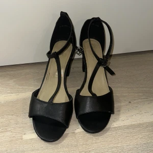 Svarta klackade sandaler med T-rem - Stilrena svarta sandaler med T-rem och öppen tå. Skorna har en bred rem över tårna, justerbart spänne vid ankeln och en stabil hög klack. Perfekta för dig som vill ha en klassisk look med lite edge. Tillverkade i syntetmaterial.