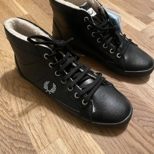 Svarta sneakers från Fred Perry, stl 37 - Svarta höga sneakers från Fred Perry med vit logga på sidan. Skorna har snörning, rund tå och är fodrade med mjukt, ljust teddyfoder för extra värme. Ovandel i skinn och slitstark gummisula. Perfekta för kyligare dagar och snygg streetstil. Helt nya skor som sticker ut på vintern.