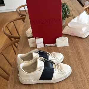 Valentino Garavani Open sneakers vit/blå - Snygga Valentino Garavani Open sneakers i vitt skinn med bred marinblå rem över vristen och coola nitar på hälen. Klassisk rund tå, platt sula och vita blå snören. Perfekta för dig som vill ha en lyxig och stilren look med edge.