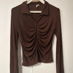 Brun rynkad topp med knappar - Snygg brun rynkad topp med knappar och med långa ärmar och krage. Framtill har den en cool rynkdetalj som ger en unik look. Perfekt för dig som gillar trendiga och bekväma plagg med lite extra attityd. Storlek M.