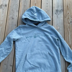 Ljusblå hoodie från Sail Racing - Ljusblå hoodie från Sail Racing med stor magficka och tryckt logga på bröstet. Tröjan har huva, långa ärmar och är gjord i mjuk bomullsmix. Perfekt för chill dagar eller när du vill ha en clean och sportig look. Dm för köp