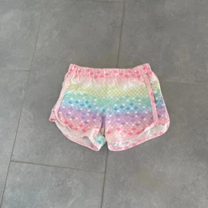 Pastellfärgade shorts med fjällmönster - Säljer ett par färgglada shorts med regnbågsfärgade fjällmönster i pastell. De har elastisk midja och är perfekta för varma sommardagar. Shortsen har en loose passform och är superbekväma för chill eller aktivitet.