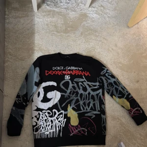 Dolce & Gabbana svart graffiti tröja - Säljer en svart långärmad tröja från Dolce & Gabbana med unikt graffitiinspirerat tryck i vitt, rött, grått, gult och rosa. Tröjan har rund hals och cool streetstyle-vibe, perfekt för dig som vill sticka ut med en statement piece.