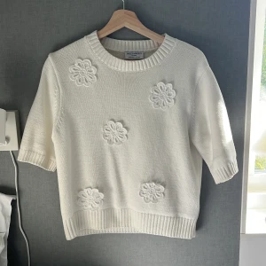 Vit stickad tröja med 5 blommor på framsidan Holly & Whyte - Superfin vit stickad tröja från Holly & Whyte med korta ärmar och broderade blommor framtill. Tröjan har ribbade muddar vid hals, ärmslut och nederkant. Perfekt för vår och sommar när du vill ha en söt och stilren look.