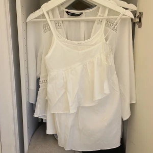 Vit volang-linnetopp från Zara - Supersöt vit linnetopp från Zara med tunna axelband och volangdetaljer framtill. Luftig och flowy känsla, perfekt för varma dagar. Tillverkad i ett lätt material som ger en snygg siluett. Enkel att styla med jeans eller kjol.