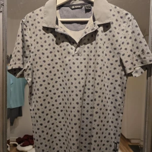 Grå paisleymönstrad pikétröja Abacus - Snygg grå pikétröja från Abacus med mörkblå paisleymönster över hela tröjan. Klassisk krage och korta ärmar, perfekt för en stilren och avslappnad look. Skön bomullskänsla och passar dig som gillar detaljer.