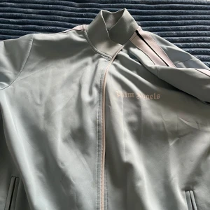 Ljusblå vindjacka Palm Angels - Säljer en ljusblå vindjacka från Palm Angels med dragkedja framtill och vita ränder med svart detalj längs ärmarna. Jackan har hög krage och broderad logga på bröstet. Perfekt för dig som gillar streetwear och snygga detaljer.