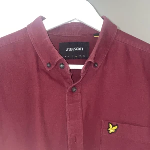 Vinröd skjorta  Lyle & Scott - Vinröd skjorta från Lyle & Scott med klassisk button down-krage och svart knappstängning. Storlek L men passar M