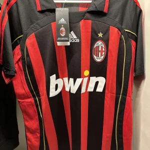 AC Milan Adidas fotbollströja randig - AC Milan matchtröja från Adidas med klassiska röda och svarta vertikala ränder, klubbmärke och guldfärgad stjärna på bröstet. Kortärmad modell i lätt och ventilerande material, med Bwin-logga på magen och detaljer i gult längs sidorna.