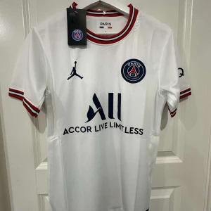 PSG vit matchtröja Jordan x Nike - Vit PSG fotbollströja med korta ärmar, tillverkad i lätt och ventilerande material. Tröjan har röda och marinblå ränder på krage och ärmslut, PSG-emblem på bröstet, Jordan-logga och stor sponsorlogga framtill. Perfekt för dig som älskar Paris Saint-Germain.