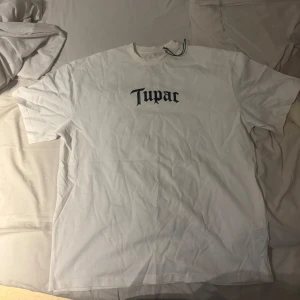 Vit Tupac t-shirt från Pull&Bear - Oversize Vit t-shirt från Pull&Bear med stort svart Tupac-tryck på bröstet och en stor bild av Tupac på ryggen. Klassisk rund hals och korta ärmar. Skön bomullskvalitet och loose fit, perfekt för dig som gillar streetstyle och hiphop-vibe.