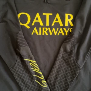 PSG x Jordan långärmad träningströja - Svart långärmad PSG träningströja från Jordan med gul logga, Qatar Airways-tryck och halv dragkedja. Ärmarna har diskret rutmönster och tröjan är i slim fit-modell. Materialet är lätt och andas, perfekt för fotbollsträning eller chill.