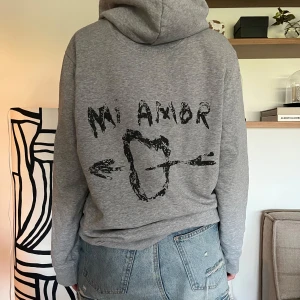 Grå hoodie med tryck 'Mi Amor' - Grå hoodie med svart tryck på ryggen där det står 'Mi Amor' och ett hjärta med en pil. Köpt på Zalando för 1700kr, märket är Mira Paris
