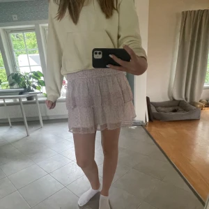 Ljusrosa volangkjol med blommigt mönster - Supersöt ljusrosa kortkjol med småblommigt mönster och volanger. Kjolen har resår i midjan för skön passform och är gjord i ett tunt, luftigt material som känns perfekt för varmare dagar.