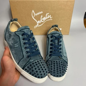 Blå mocka sneakers från Louboutin - Unika blå sneakers från Christian Louboutin i mjuk mocka med coola nitar på tån. Skorna har mörkblå snörning, vit sula och rund tå. Perfekta för dig som vill sticka ut med en edgy och lyxig stil.