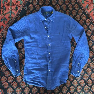 Blå linneskjorta från Ralph Lauren XS - Snygg blå skjorta från Ralph Lauren i slim fit-modell. Tillverkad i linne med klassisk krage, vita knappar och broderad logga på bröstet. Perfekt för dig som gillar stilrena och bekväma plagg med en exklusiv touch.