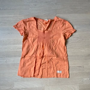 Orange blus från Odd Molly - Superfin orange blus från Odd Molly med virkade detaljer vid halsringningen och smock framtill. Blusen har korta ärmar med söt kant och är gjord i tunn bomull, perfekt för varma dagar. Liten broderad Odd Molly-logga nertill.
