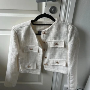 Vit kavajjacka med guldknappar - Snygg vit kavajjacka från ZARA i strukturerat tyg med fyra fickor framtill och dekorativa guldknappar. Jackan är croppad och har lång ärm, perfekt för en chic och trendig look.