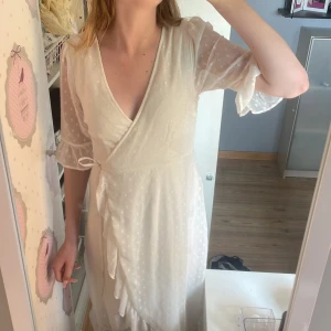 Vit omlottklänning med prickigt mönster - Superfin vit långklänning med omlottmodell och knyt i midjan. Klänningen har transparenta trekvartsärmar med volangkant och ett diskret prickigt mönster i tyget. Perfekt för sommarens alla tillfällen!