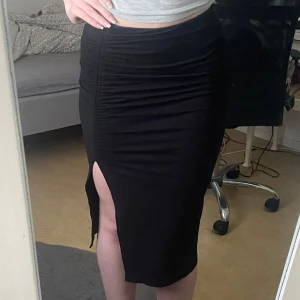 Svart långkjol med slits från H&M Divided - Svart långkjol från H&M Divided i figurnära modell med snygg slits framtill. Kjolen har rynkade detaljer längs ena sidan som ger en cool look. Perfekt för dig som gillar stilrena plagg med en twist. Använd ett fåtal gånger💕