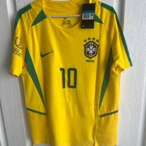 Brasilien Rivaldo 10 fotbollströja Nike - Gul Brasilien fotbollströja från Nike med gröna detaljer, nummer 10 och Rivaldo på ryggen. CBF-emblem och VM 2002-logga på ärmen. Klassisk rund hals och korta ärmar. Perfekt för dig som älskar fotboll och vill hylla en legend.