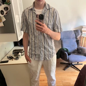 Randig kortärmad skjorta från Hugo Boss - Säljer en snygg kortärmad skjorta från Hugo i storlek M. Skjortan har ett randigt mönster i blått, vitt och grått, klassisk krage och knäppning framtill. Perfekt för dig som gillar en clean och fräsch stil med lite edge.