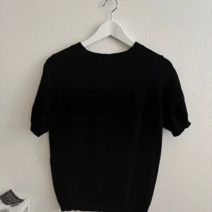 Svart stickad kortärmad tröja - Enkel och stilren svart stickad tröja med korta ärmar och rund halsringning. Perfekt för dig som gillar minimalistisk stil och vill ha något som funkar till många olika outfits. Mjuk och skön känsla tack vare det stickade materialet.