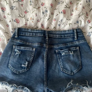 Blå jeansshorts med slitningar - Snygga blå jeansshorts med slitna detaljer och fransig nederkant. Klassisk femficksmodell med bälteshällor och dragkedja fram. Perfekta för en avslappnad och trendig look under varma dagar.