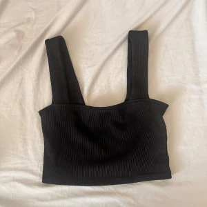 Svart ribbad linnetopp - Snygg svart ribbad linnetopp med breda axelband och croppad passform. Perfekt att styla med höga jeans eller kjol för en trendig look. Toppens ribbade material ger en cool struktur och sitter skönt mot kroppen.