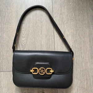 Svart axelväska från Guess  - Snygg svart axelväska från Guess i slätt skinn med guldfärgade metalldetaljer och logga framtill. Väskan har justerbart axelband med nitar, dragkedjeficka på baksidan och ljus insida med Guess-tryck. Perfekt storlek för det viktigaste.