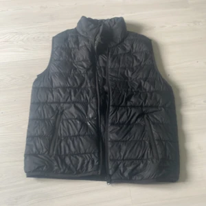 Svart dunväst från Kappahl, stl 170 - Svart quiltad dunväst från Kappahl i storlek 170. Västen har hög krage, två dragkedjefickor och stängs med dragkedja framtill. Perfekt att slänga över en hoodie för extra värme. Snygg och enkel design som funkar till det mesta.