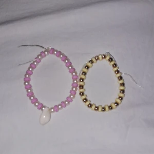 Två pärlarmband med snäcka och pärlor - Säljer två handgjorda armband med pärlor. Ett armband har rosa och vita pärlor samt en liten snäcka, det andra har gula och guldiga pärlor. Båda är elastiska och passar de flesta handleder. Perfekta för att ge din stil en somrig och personlig touch.