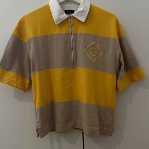 Randig pikétröja från Gant i gult och beige - Säljer en kortärmad pikétröja från Gant med breda gula och beige ränder, vit krage och halv dragkedja framtill. Tröjan har broderad G-logga på bröstet och är tillverkad i 100% bomull. Perfekt för en sportig och avslappnad look.