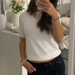 Vit ribbad topp från Zara - En stilren vit topp från Zara med ribbade detaljer i nederkant och ärmslut. Modellen har rund halsringning och korta ärmar som slutar strax ovanför armbågen. Perfekt att matcha med både jeans och kjol för en clean look.