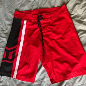 Röda Better Bodies shorts med snörning - Snygga röda shorts från Better Bodies med svart och vit rand på ena benet och stor logga. De har snörning i midjan och är tillverkade i polyester, perfekta för stranden eller träning. Sportig look och skön passform.