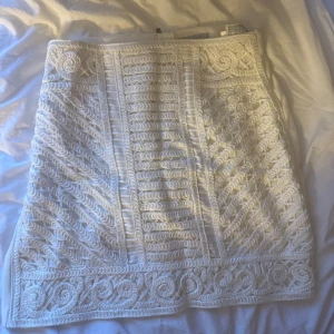 Vit kortkjol med broderade detaljer - Superfin vit kortkjol med broderade mönster och snygga strukturer framtill. Kjolen har dragkedja bak och är fodrad. Perfekt för dig som vill sticka ut med en unik och elegant look.