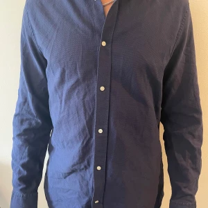 Mörkblå skjorta från J.Lindeberg - Snygg mörkblå skjorta från J.Lindeberg i klassisk modell med vita knappar och lång ärm. Skjortan är tillverkad i bomull och har en diskret struktur i tyget som ger en modern känsla. Perfekt för dig som gillar stilrena plagg med en clean look.