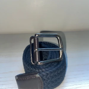 Mörkblått flätat bälte med skinn - Snyggt mörkblått bälte med flätad design och detaljer i mörkbrunt skinn. Spännet är i metall och ger en stilren känsla. Perfekt accessoar för att lyfta din outfit med en klassisk touch.